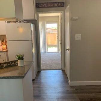 Bright kitchen with white fridge, island, gray floors, and a hallway leading to a patio door.