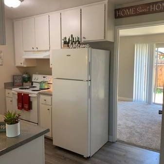Bright kitchen with white cabinets, stove, refrigerator, and a doorway to a carpeted living room.