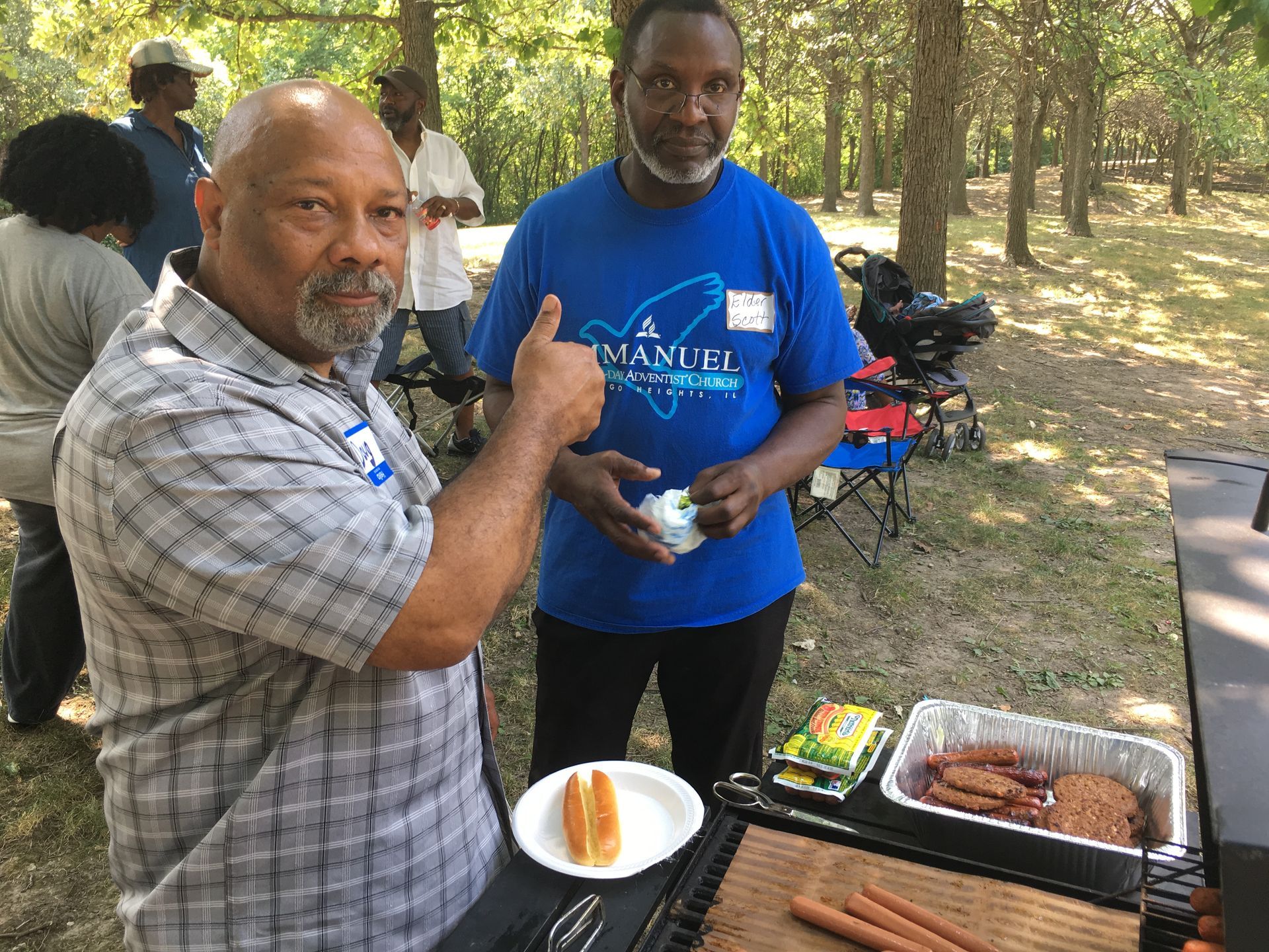 Two men at a picnic grill, one pointing at the other holding food outdoors by the woods
