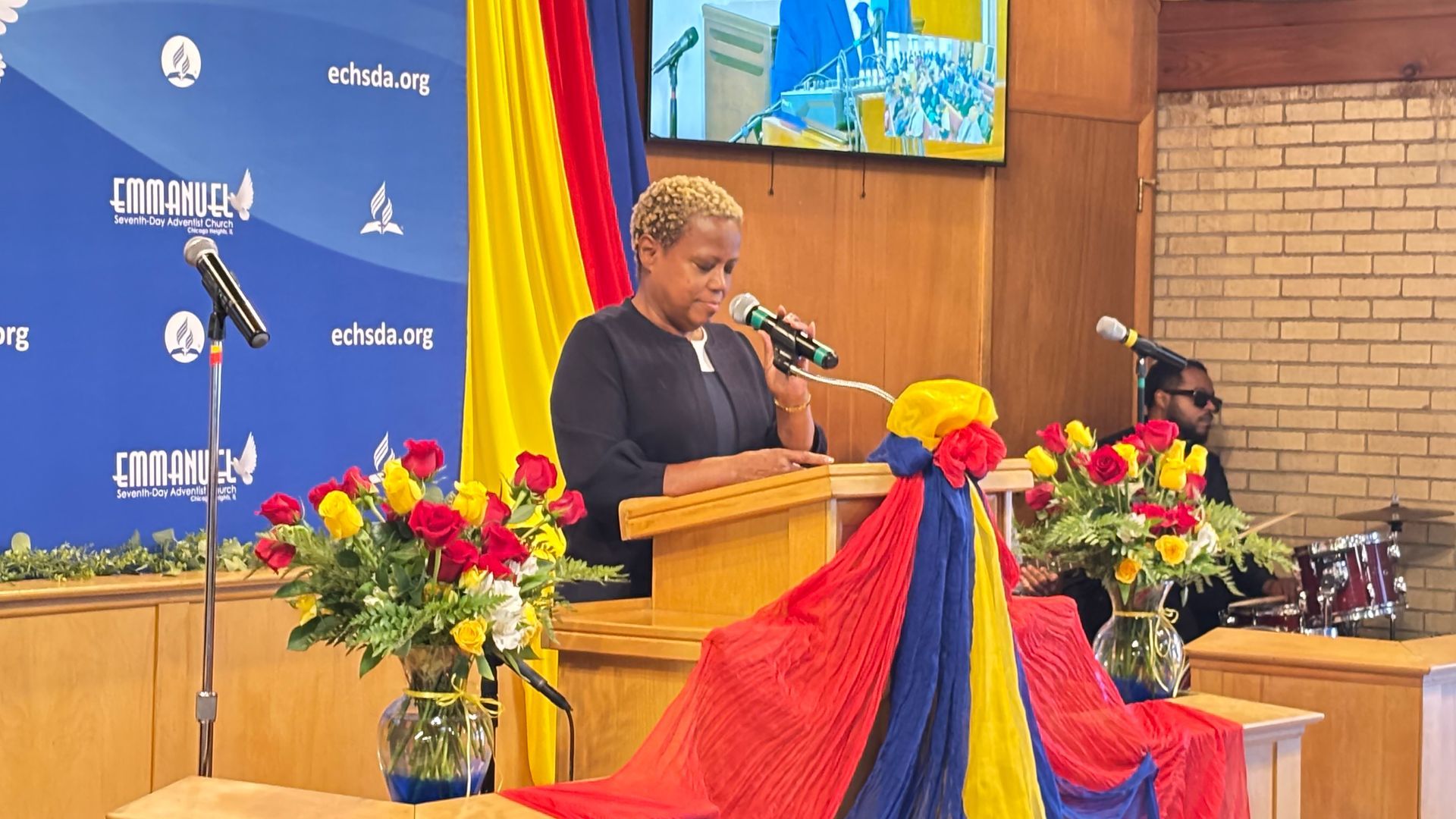 Person speaking at a podium decorated with red and yellow flowers in a church or hall.