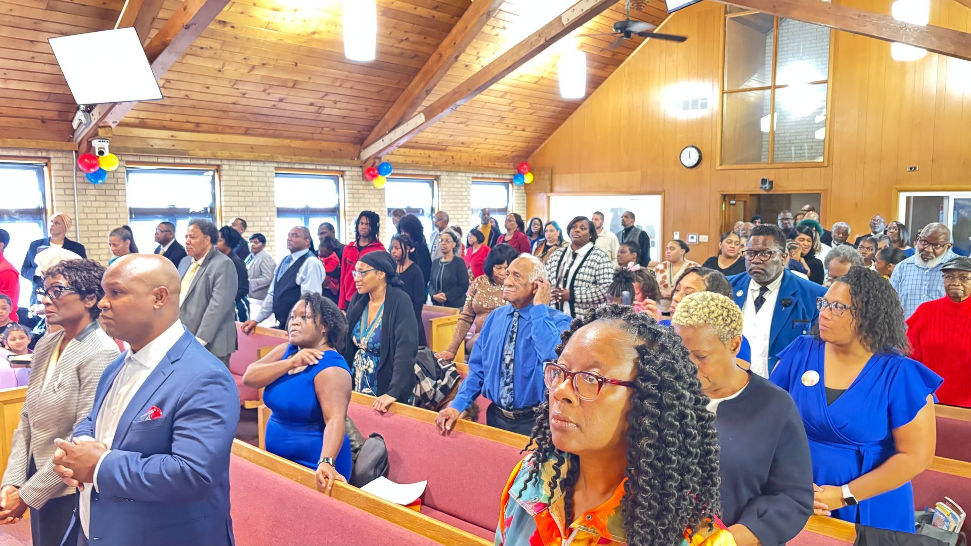 Congregation standing in a church sanctuary with wooden ceiling, red pews, and stained glass windows