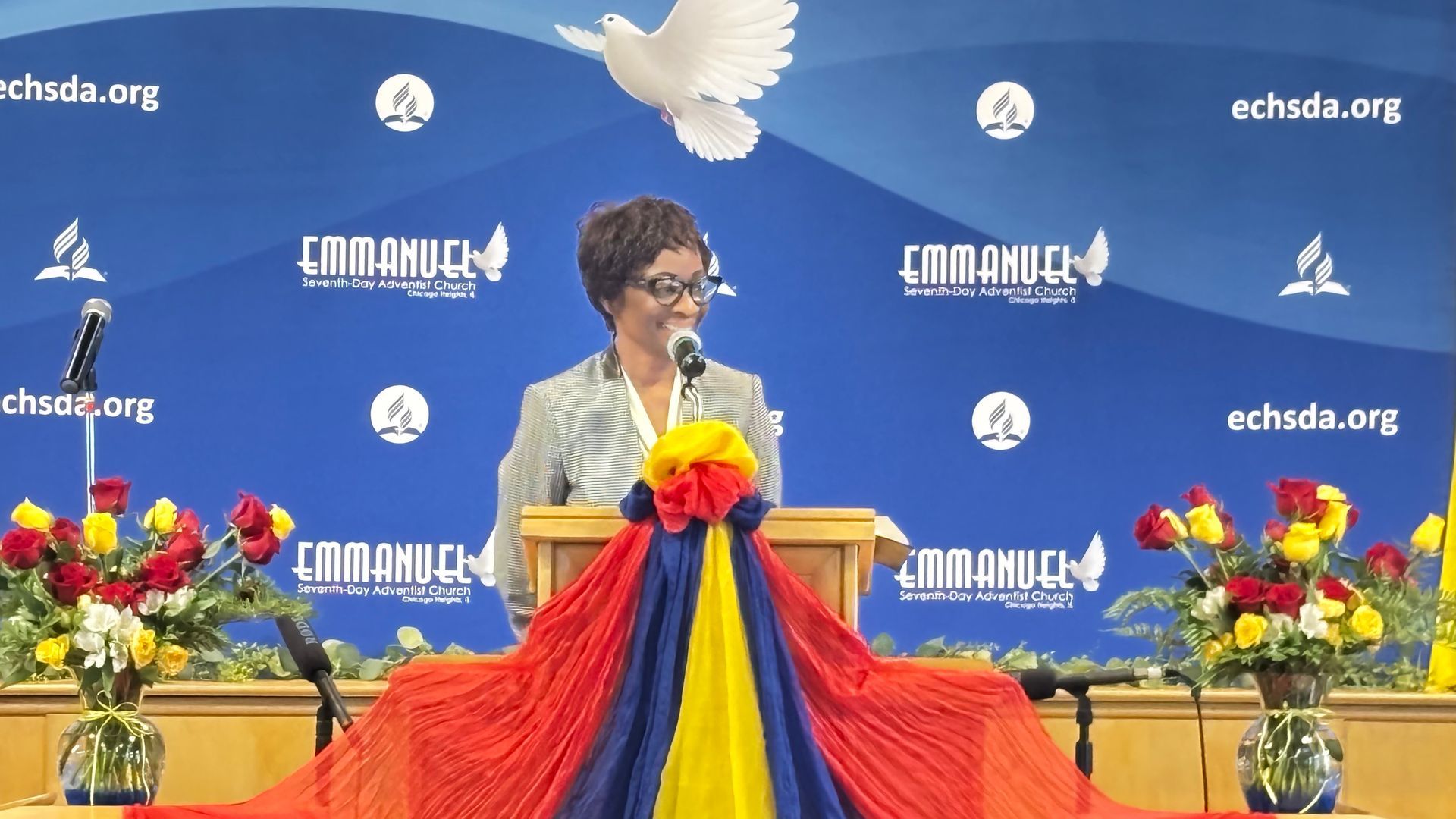 Speaker at podium with colorful draped cloth, flowers, and blue event backdrop with logos