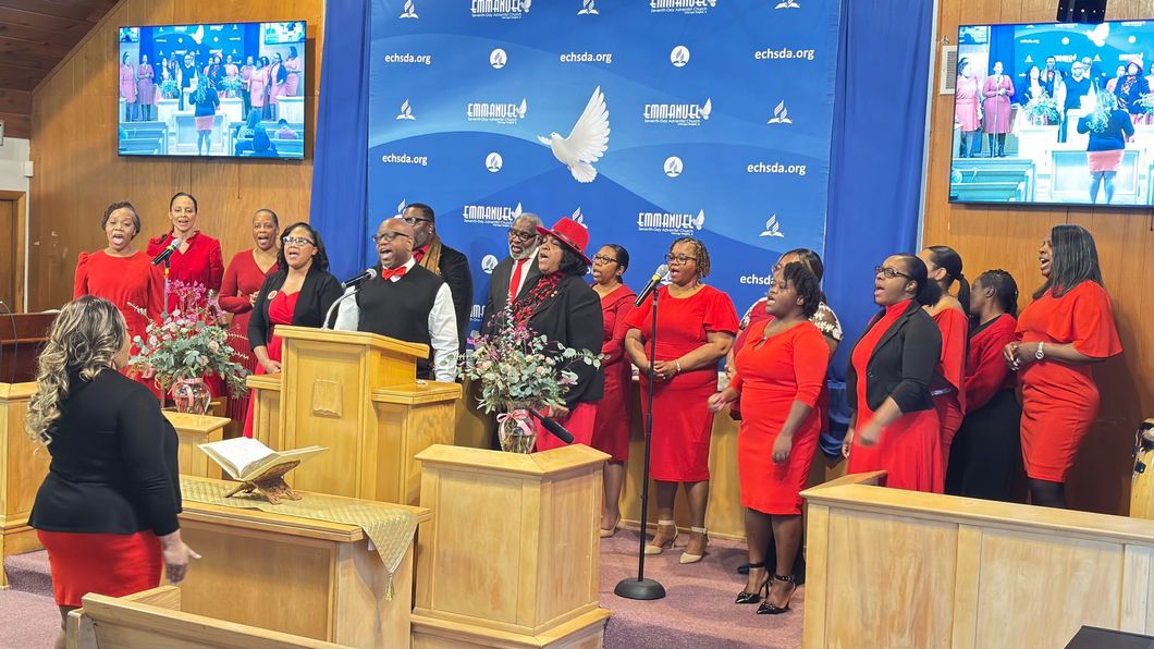 Choir in red stands beside a pulpit on a church stage with blue backdrop and screens showing the service.