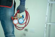 Worker holding red coiled hoses with blue and red gauges, wearing gloves and jeans indoors