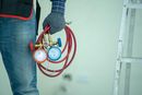 Worker holding red coiled hoses with blue and red gauges, wearing gloves and jeans indoors