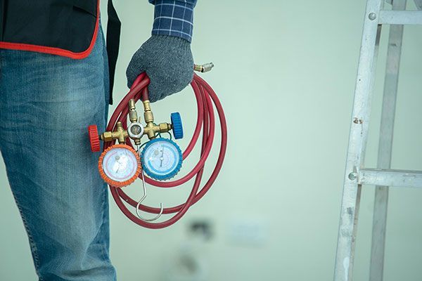 Worker holding red coiled hoses with blue and red gauges, wearing gloves and jeans indoors