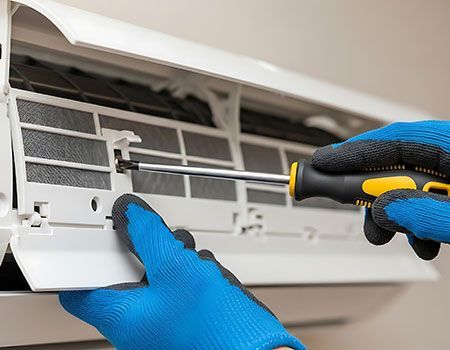 Blue-gloved hands cleaning an air conditioner filter with a screwdriver attached tool