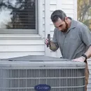 Man inspecting a rooftop HVAC unit with a tool outside a house