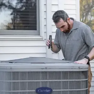 Man inspecting a rooftop HVAC unit with a tool outside a house