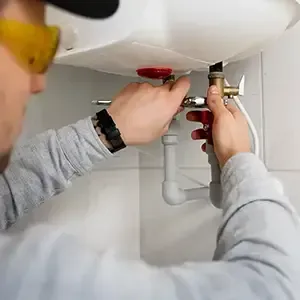 Person tightening a sink pipe under a white bathroom sink with tools, wearing safety glasses