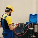 Worker in yellow shirt and blue overalls using a hose on outdoor HVAC equipment.
