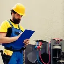 Technician in yellow hard hat and blue overalls checks a clipboard beside a machine with gauges and hoses