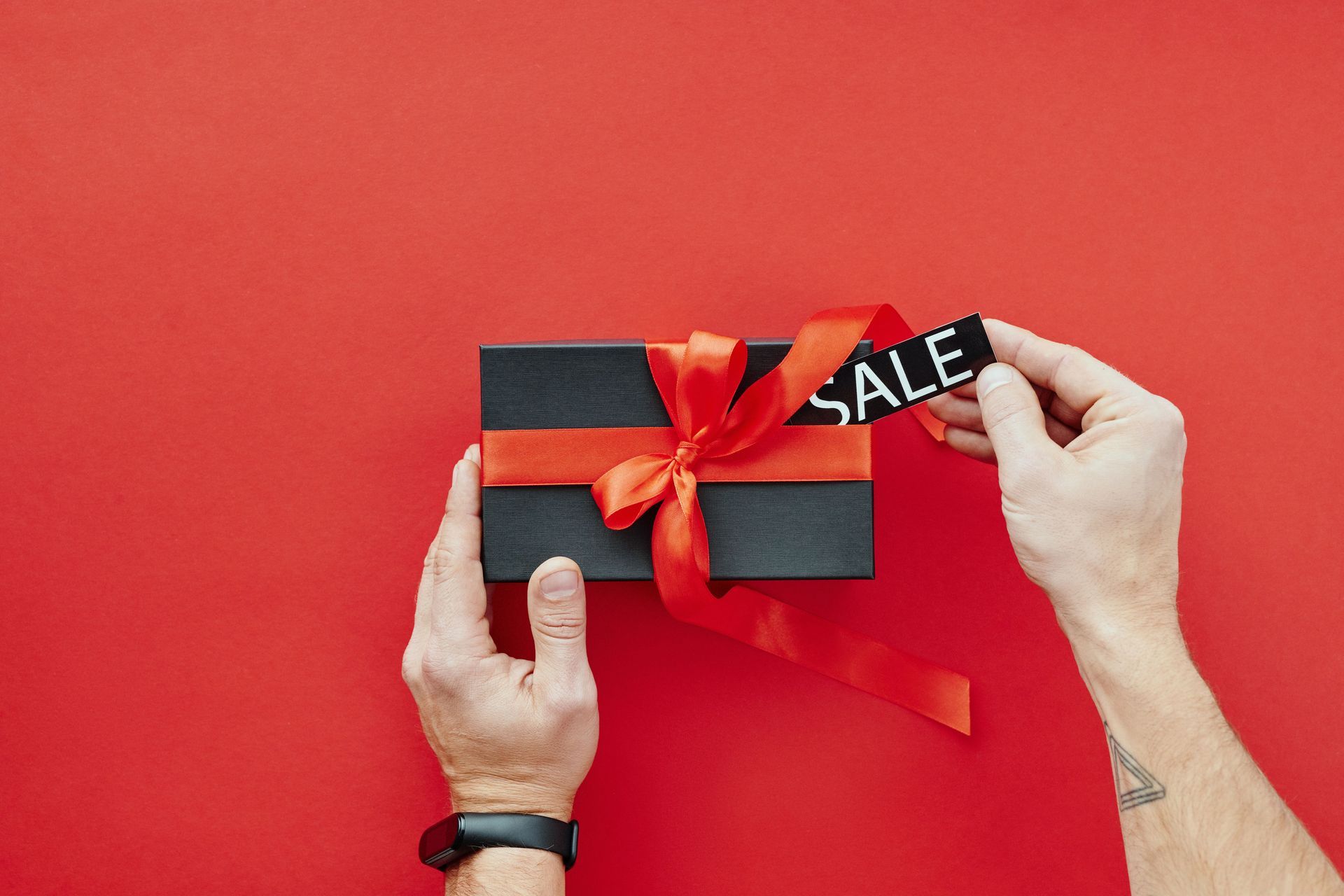 Hands holding a black gift box with a red ribbon and “SALE” tag on a red background