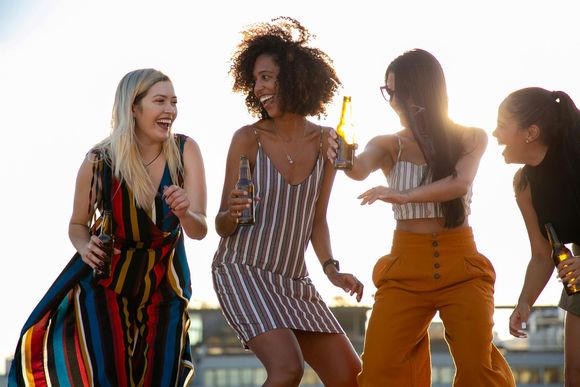 Four people laughing and dancing outdoors, holding bottles in bright sunlight.