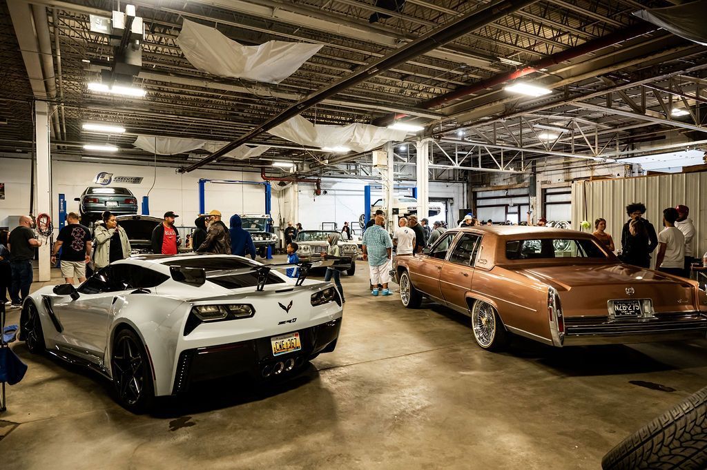 Inside car show: white Corvette and gold vintage car on display, people walking around in garage.