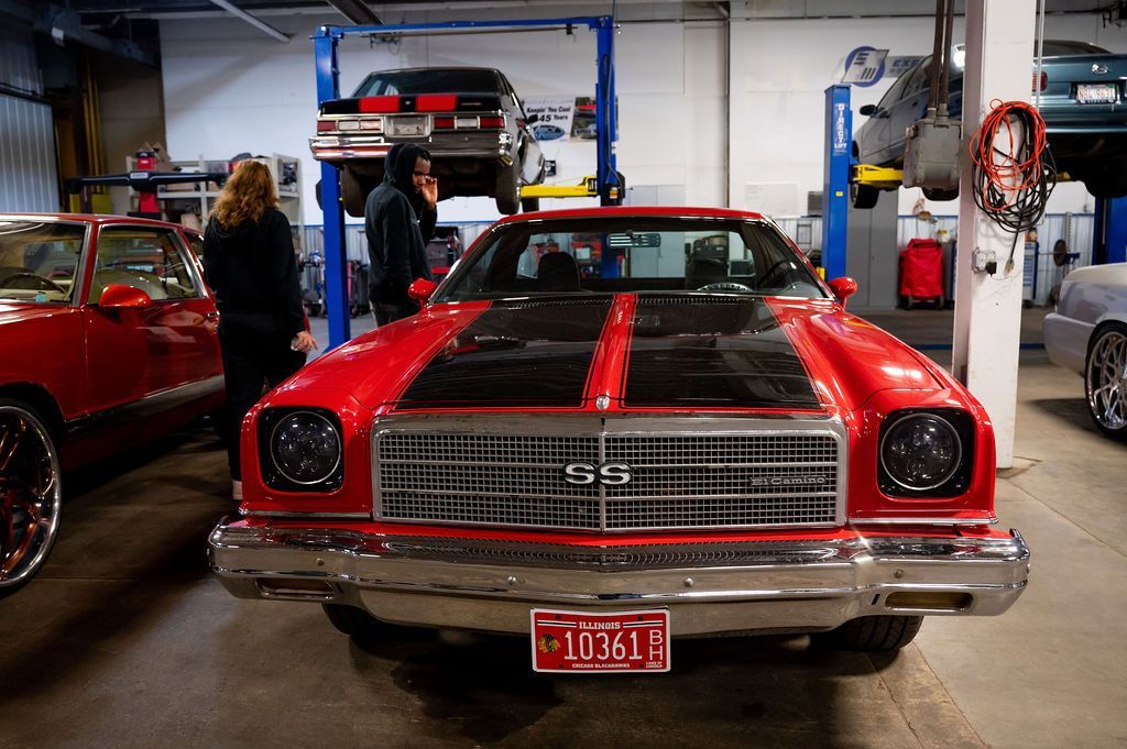 Red Chevy car with black hood stripes in a garage. People are nearby, cars on lifts.