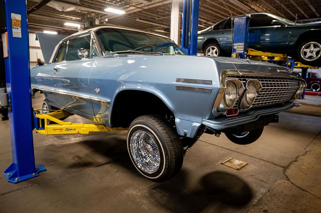 Light blue classic car on a lift in a garage, undergoing repair, with another car in the background.