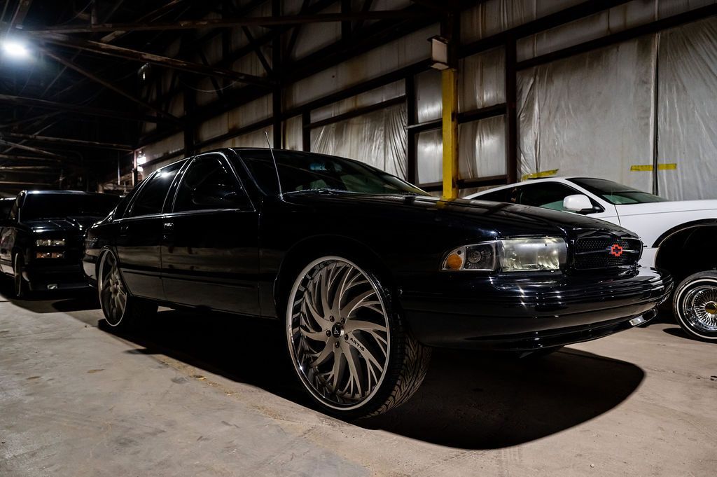 Black car with large chrome wheels parked in a dark garage, other cars are in the background.