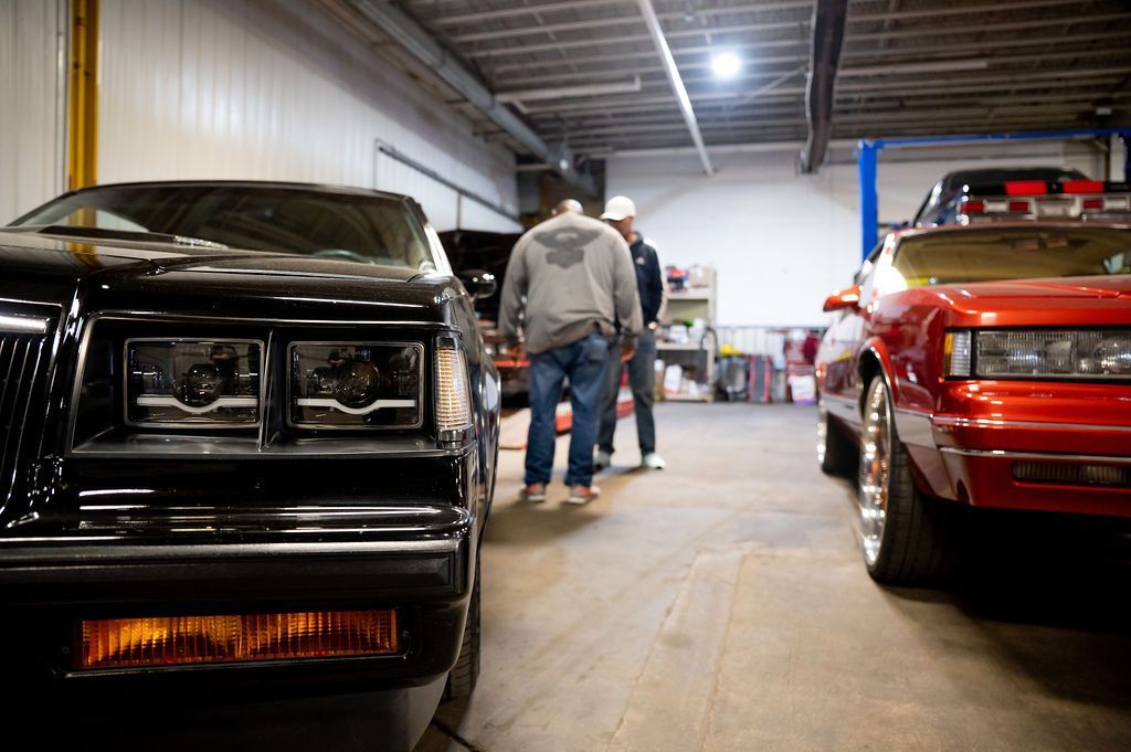 Two classic cars parked in a garage with three people walking between them.