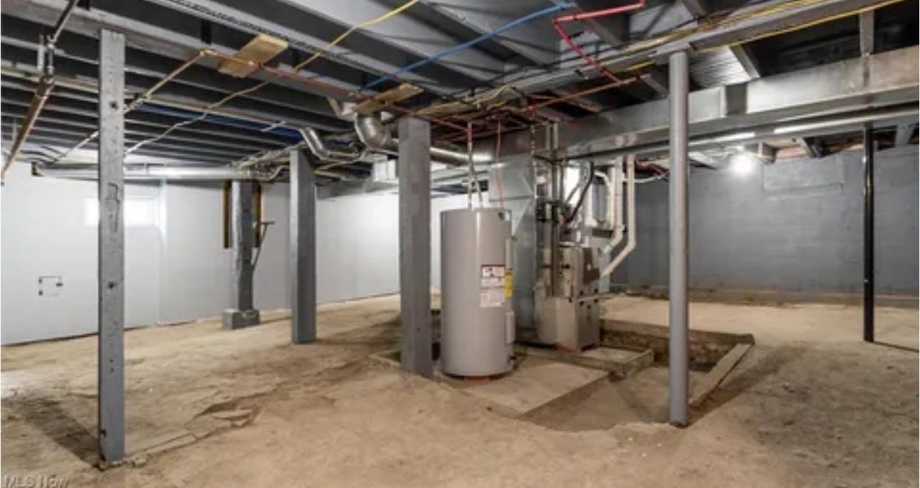 An unfinished basement with concrete floors, gray walls, support columns, a water heater, and a furnace.