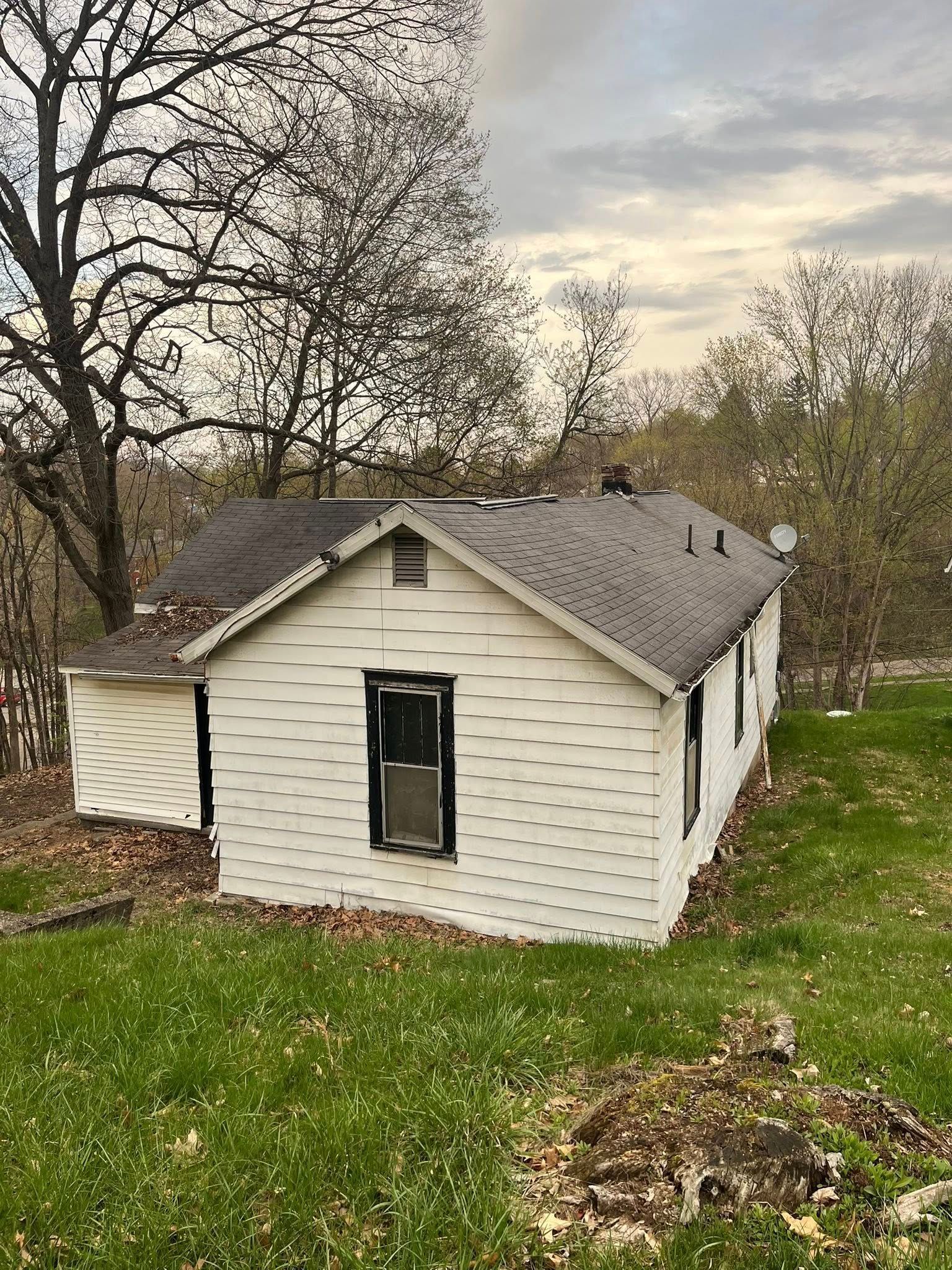 A white, single-story house with dark shingles and black-trimmed windows sits on a grassy slope beneath bare trees.