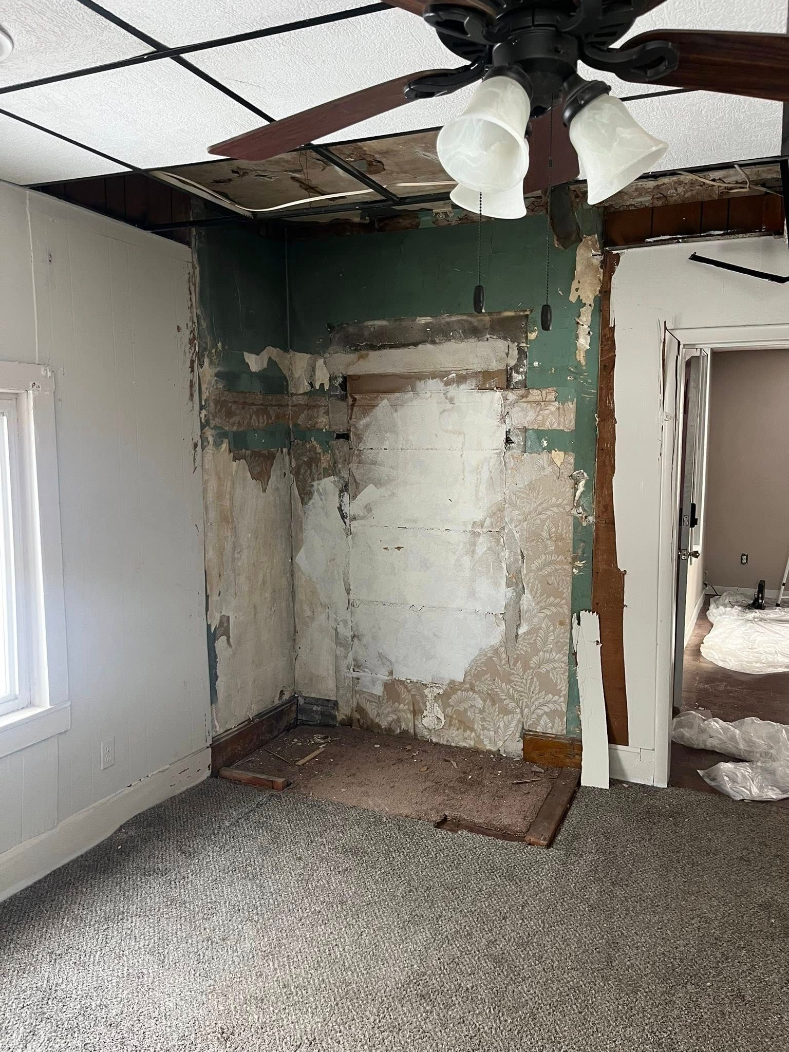 A room under renovation with exposed plaster and brick on a wall, a missing section of ceiling, and a ceiling fan.