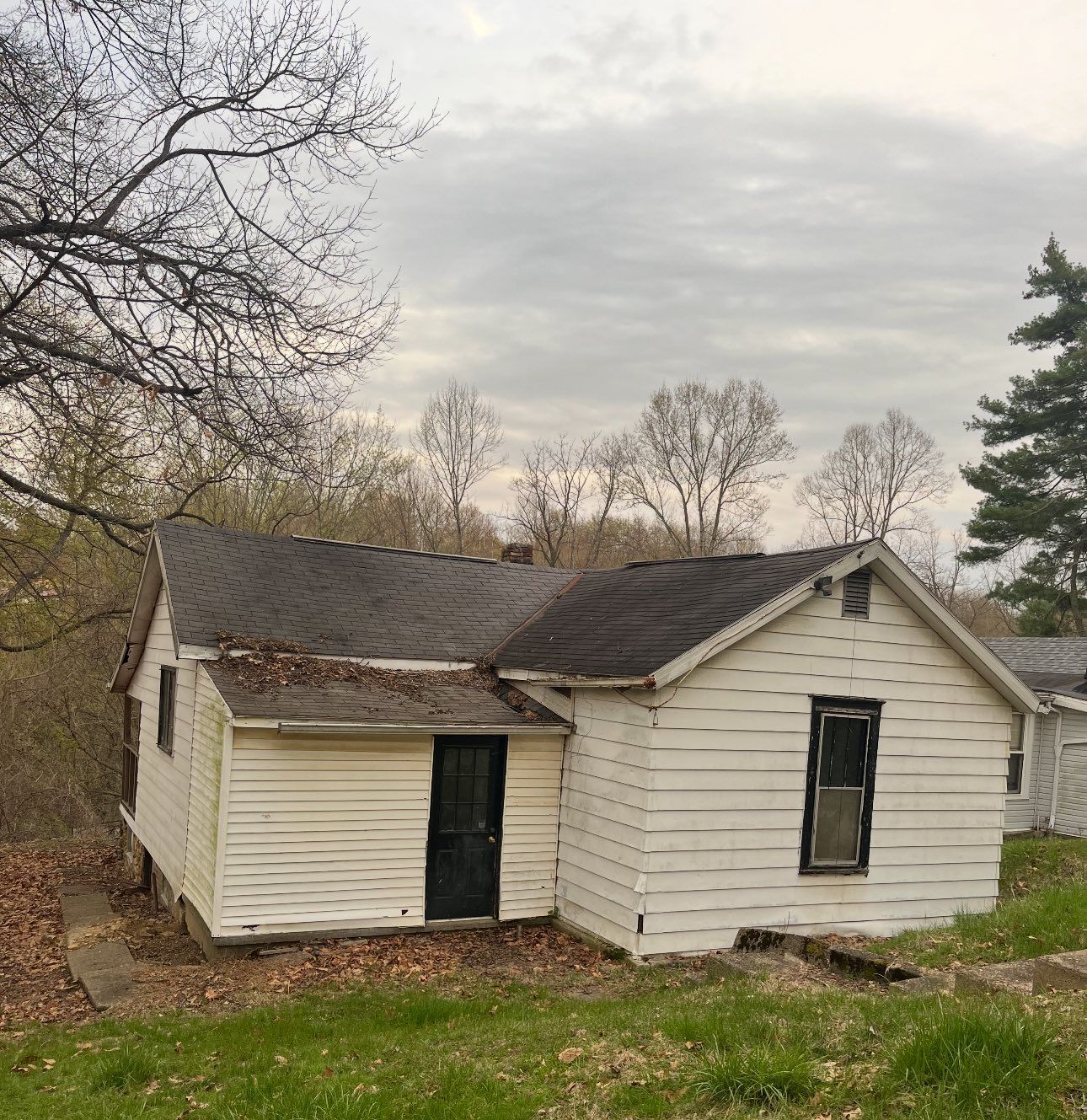 A small, weathered white cottage with a dark door and window sits on a grassy hill under a cloudy sky.