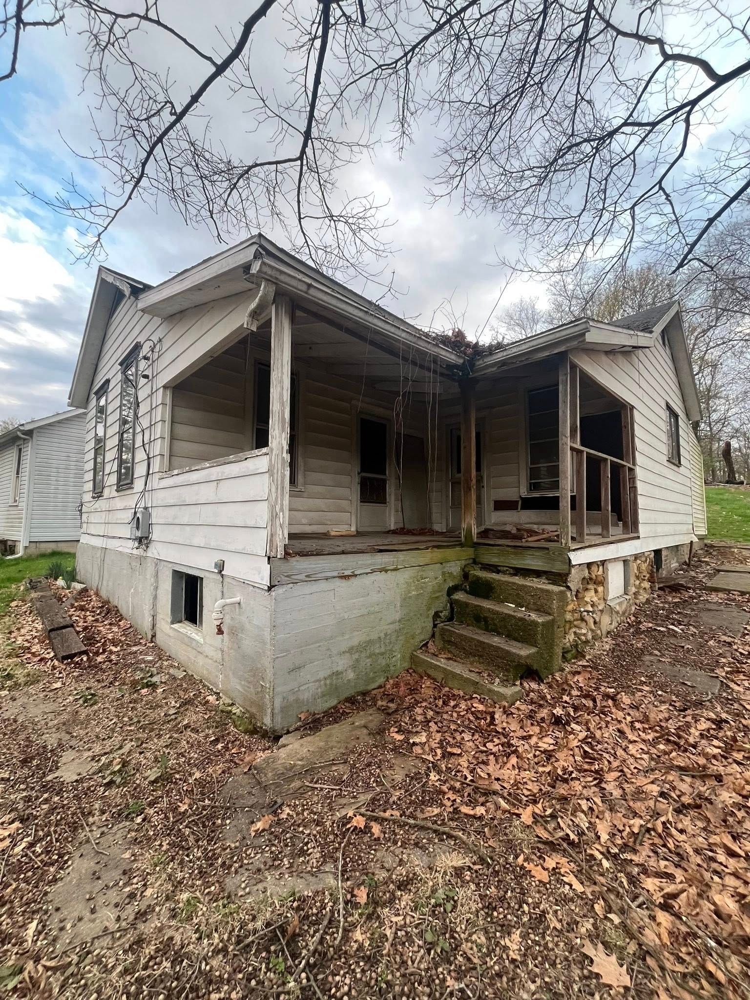 A dilapidated, white wood-frame house with a covered porch and stone foundation surrounded by fallen autumn leaves.