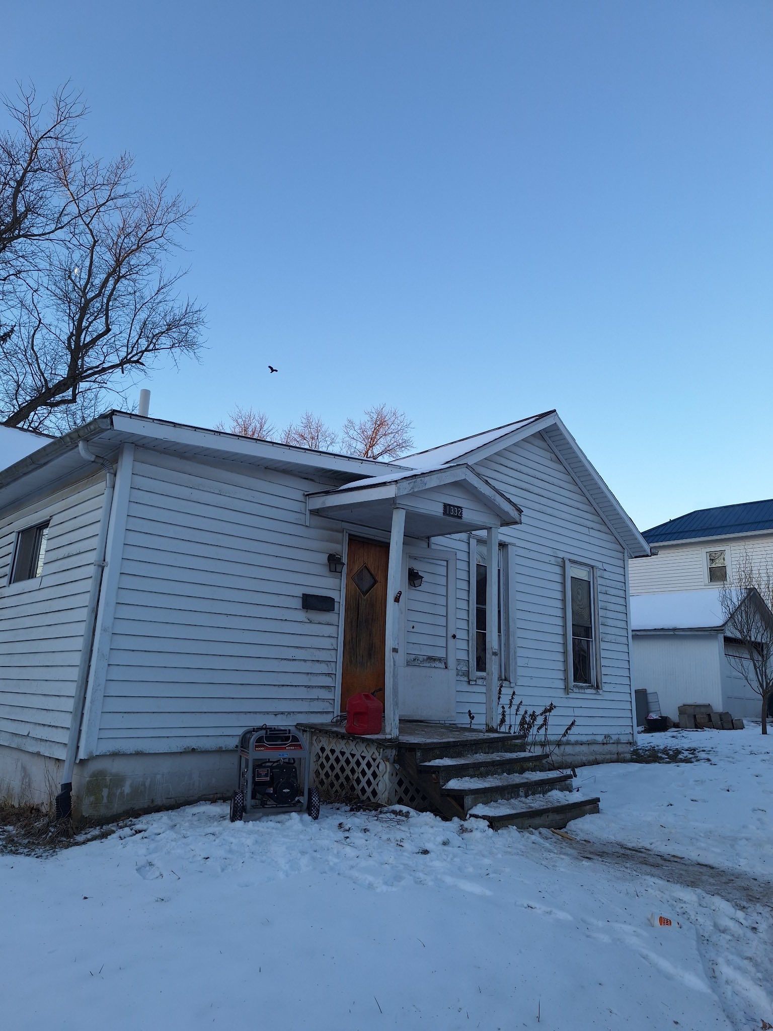 A white, single-story house with a wooden door and a generator on the porch, set in a snowy landscape under a blue sky.