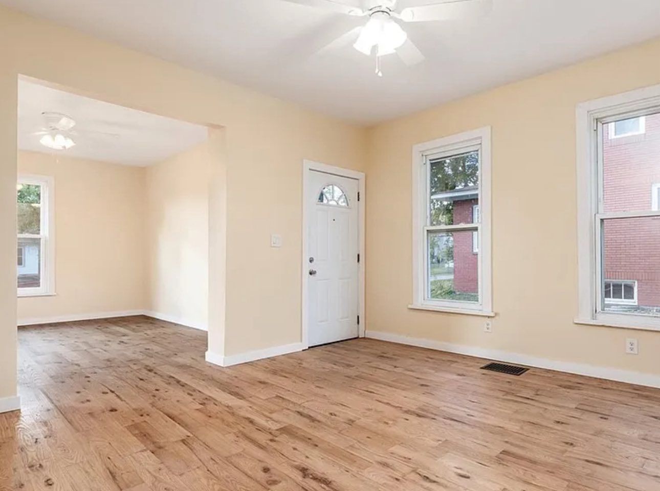 Empty room with light wood flooring, cream-colored walls, a white front door, and two windows with view of a brick house.