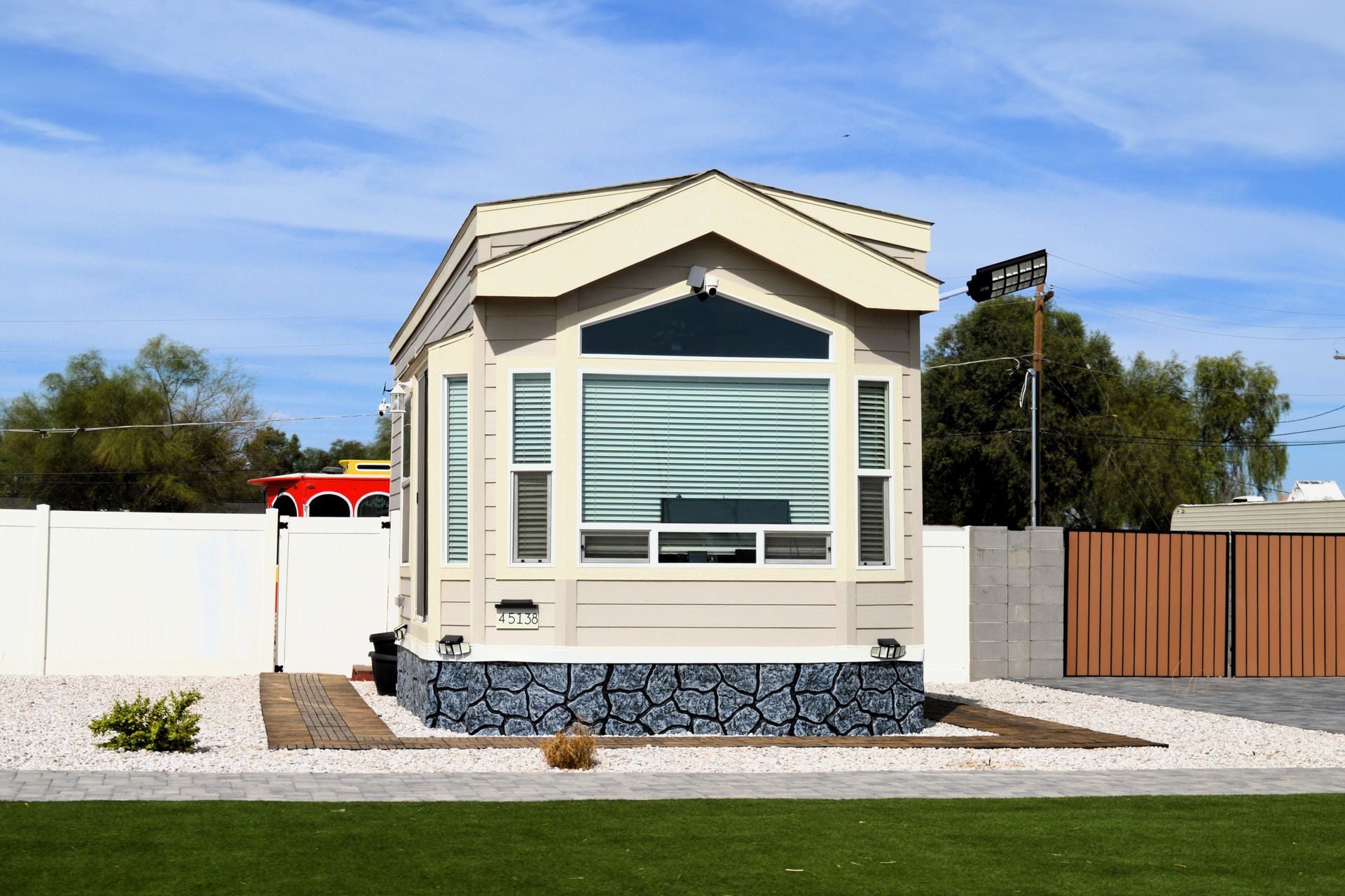 A tan-colored park model home with stone skirting, set on a gravel lot with a white vinyl fence and a blue sky above.
