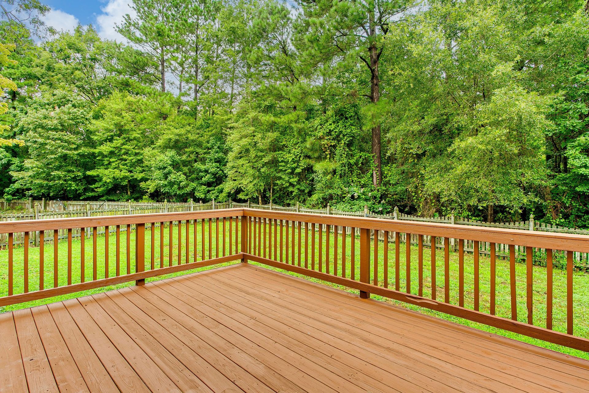 A wooden deck with a railing overlooking a grassy backyard bordered by a dense forest under a blue sky.
