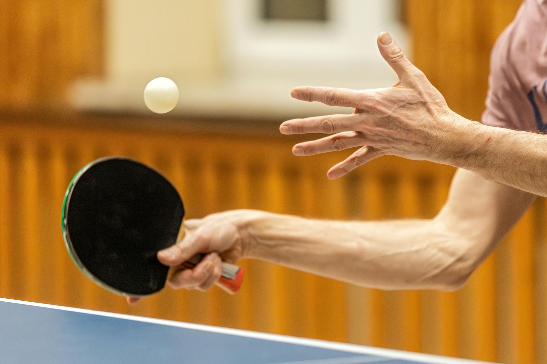 Ping pong player serving the ball with paddle and hand reaching out; indoor setting.