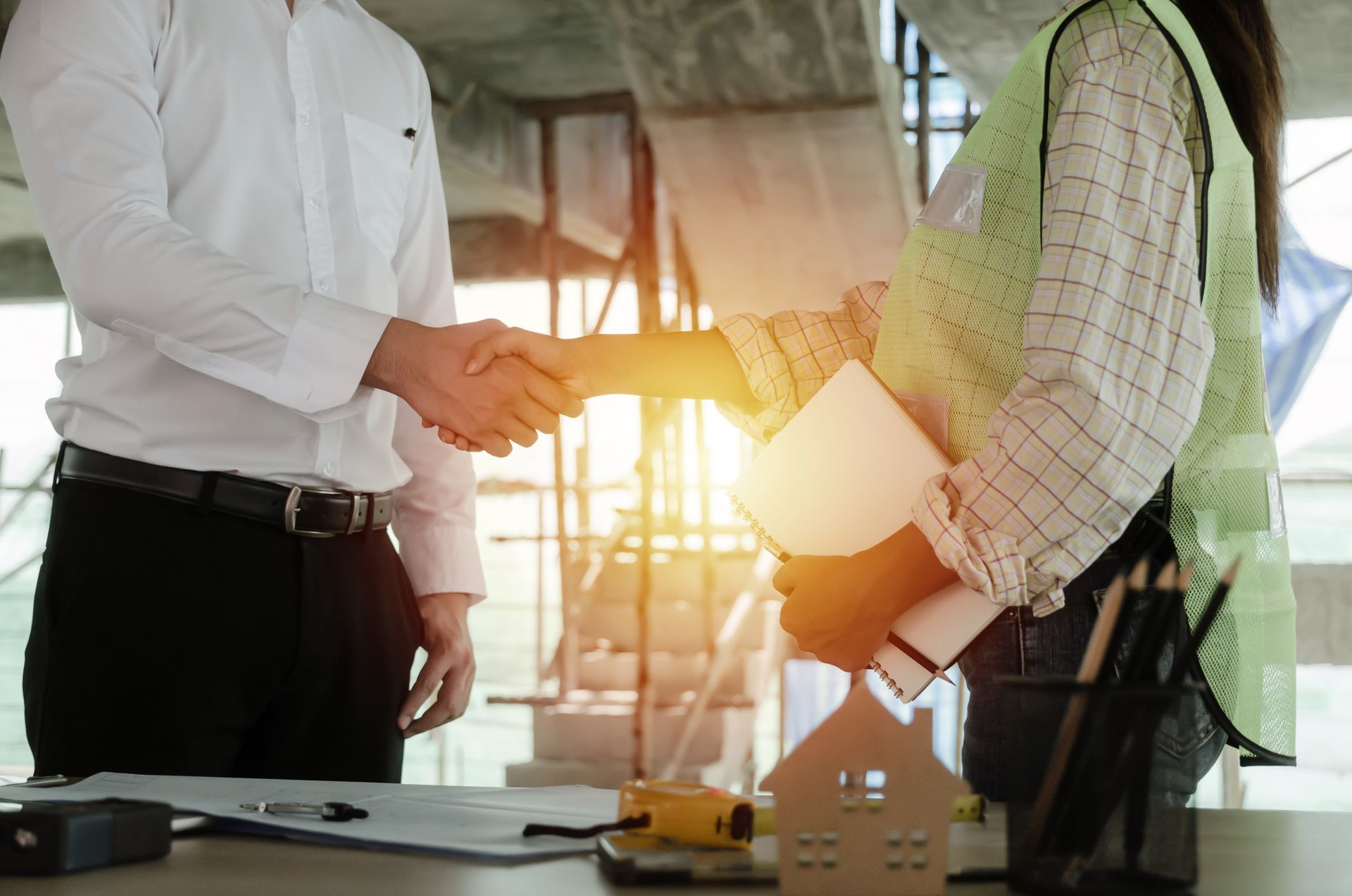 Two people shake hands across a desk in a sunlit office with a house model and blueprints. Two people shake hands across a desk in a sunlit office with a house model and blueprints.