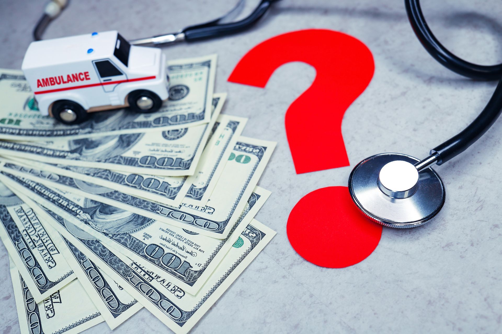 Medical bills, cash, and a stethoscope beside a red question mark on a desk Medical bills, cash, and a stethoscope beside a red question mark on a desk