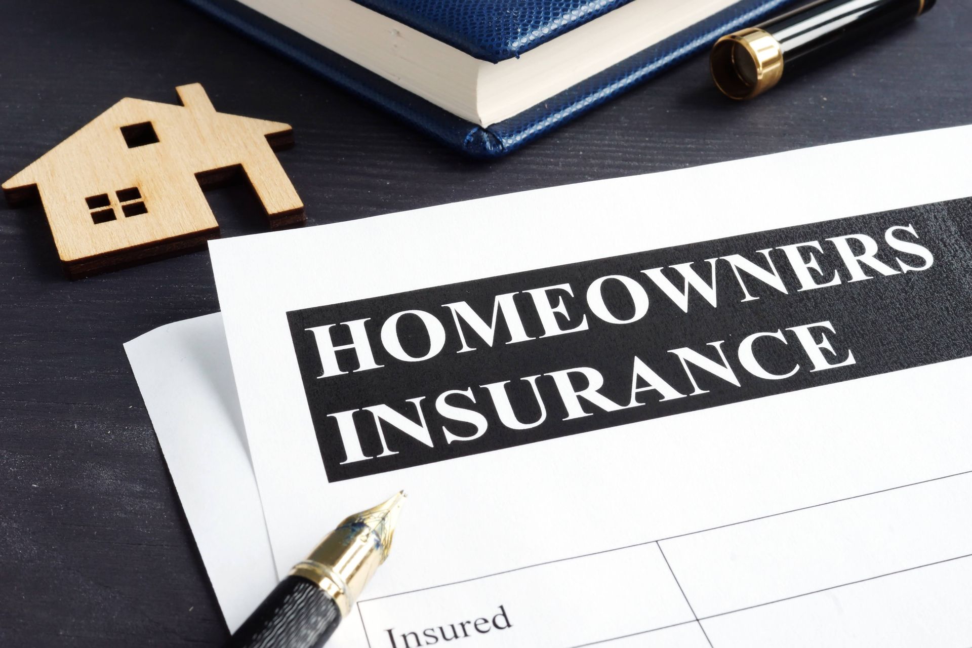 Homeowners insurance form with pen, small wooden house, and book on dark surface.