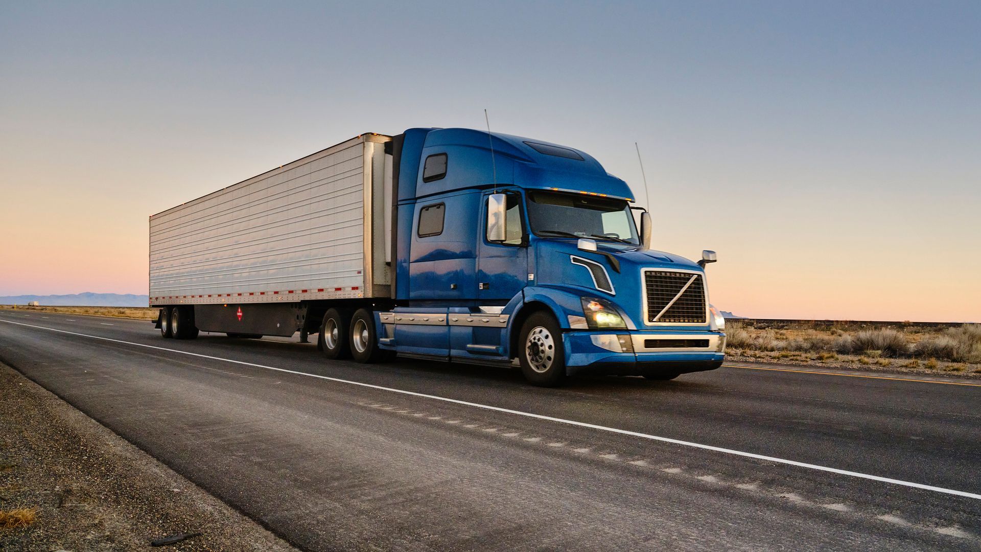 Blue Volvo semi-truck with trailer parked beside empty roadway.