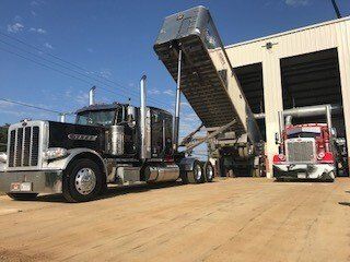 Black Peterbilt semi-truck with trailer raised, unloading at a warehouse, another red truck nearby.