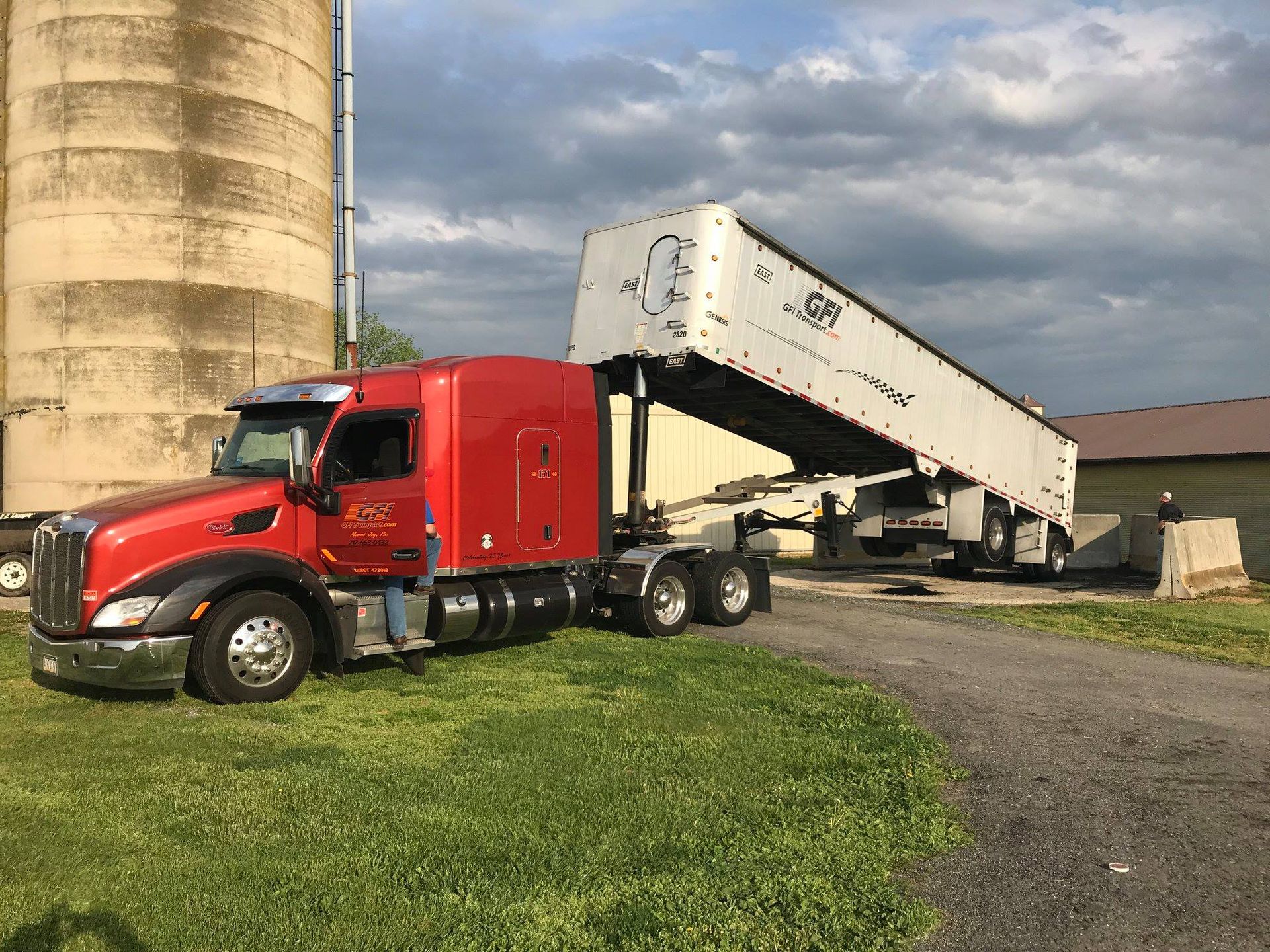 Red semi-truck hauling a white dump trailer beside a grain silo on a farm
