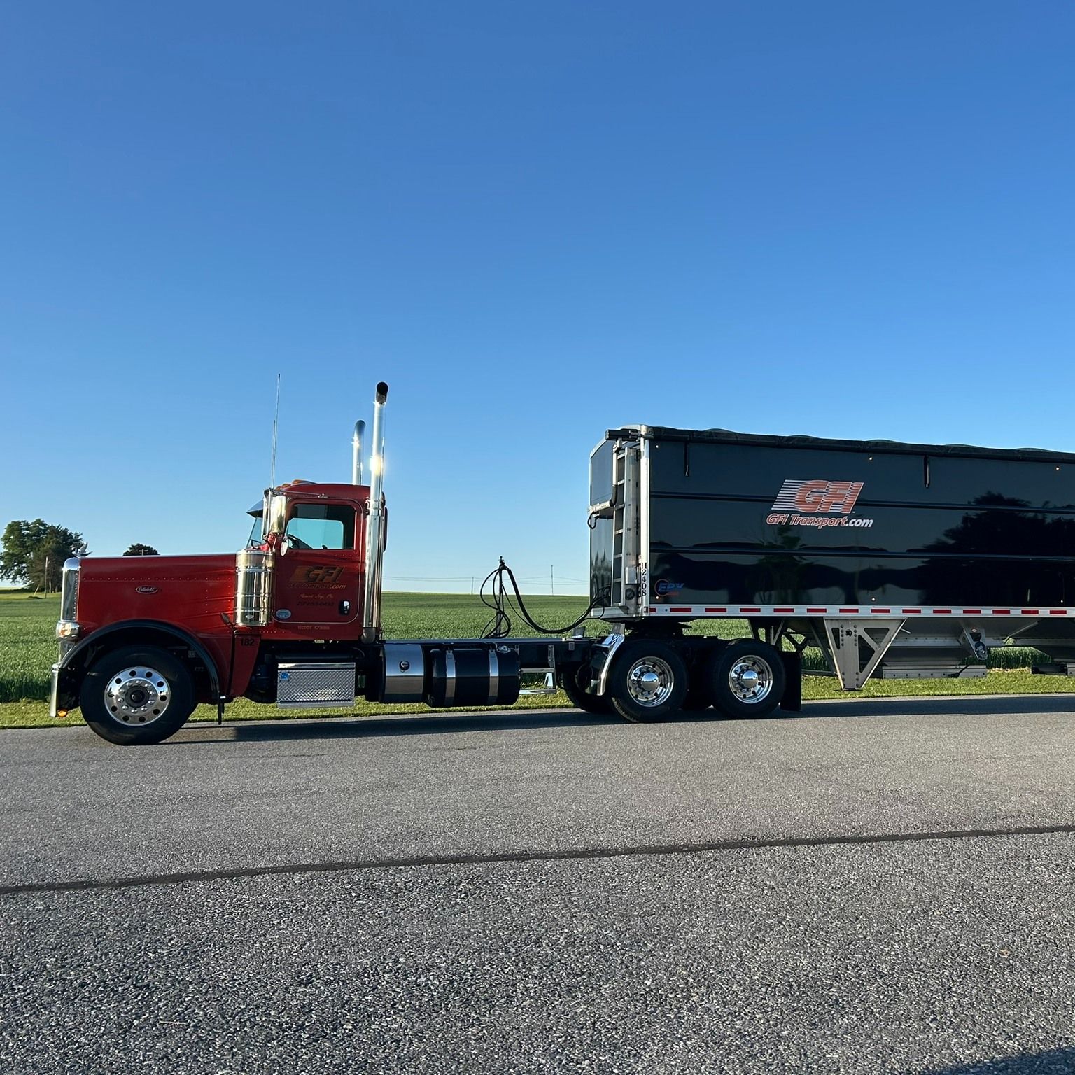 Red semi-truck with a black trailer on a road, blue sky background.