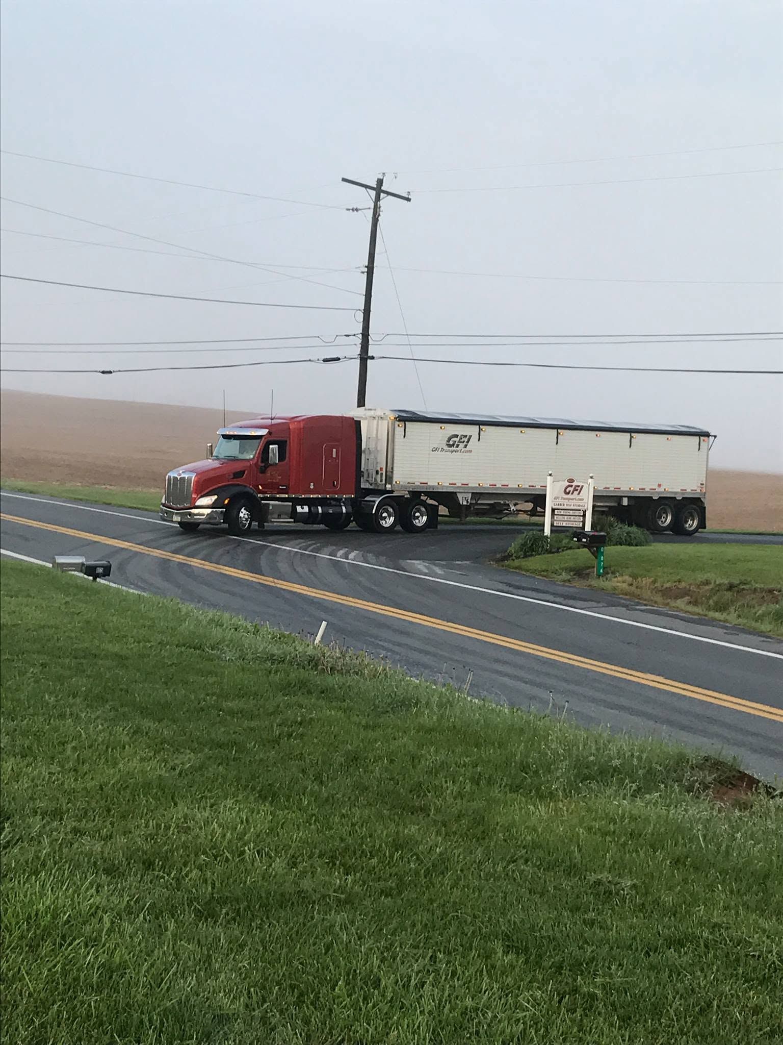 Red semi-truck with white trailer on a wet roadside beside a grassy verge and utility pole.