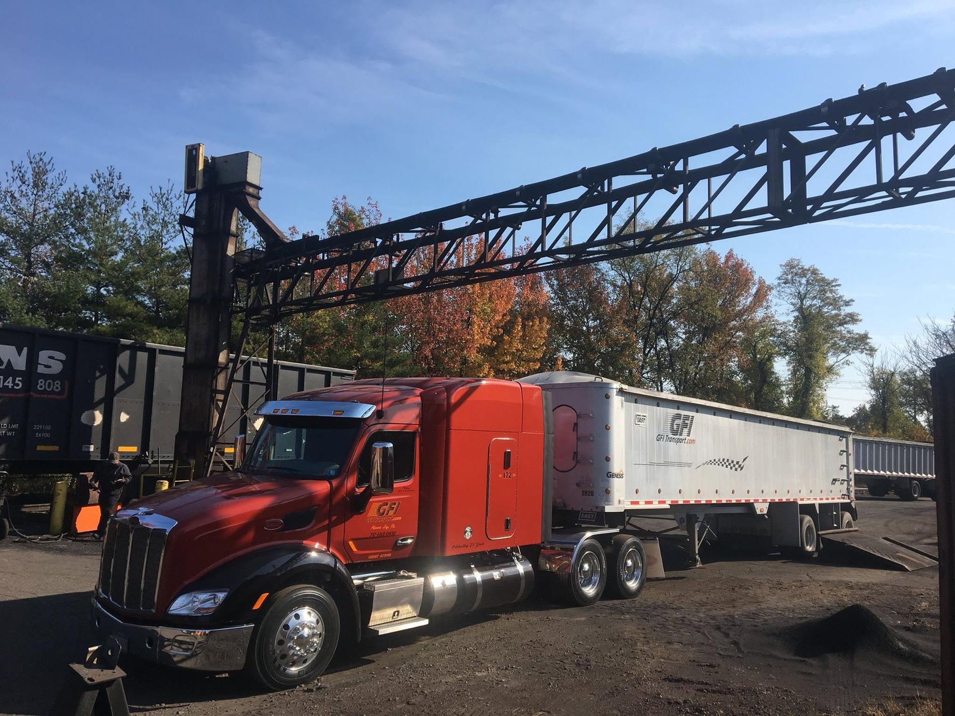 Red semi-truck hauling a white dump trailer under a conveyor in a gravel yard.