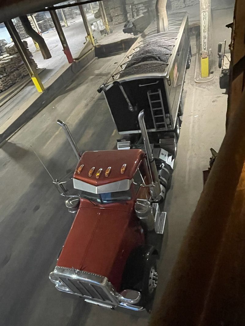 A red semi truck is parked while stone or asphalt is being loaded into the hopper bottom trailer