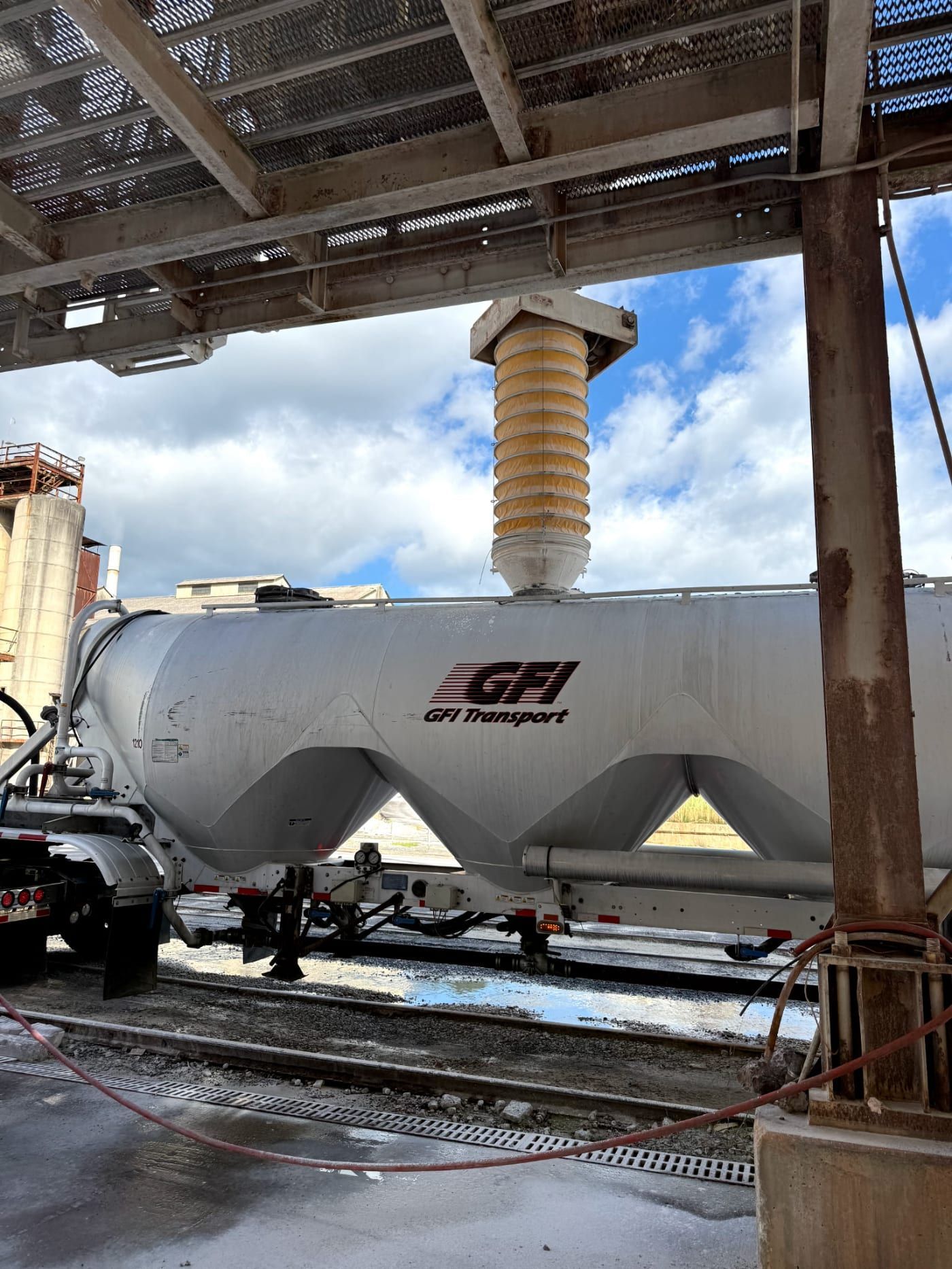 pneumatic trailer under a loading chute in an industrial shed, with metal beams and wet tracks.