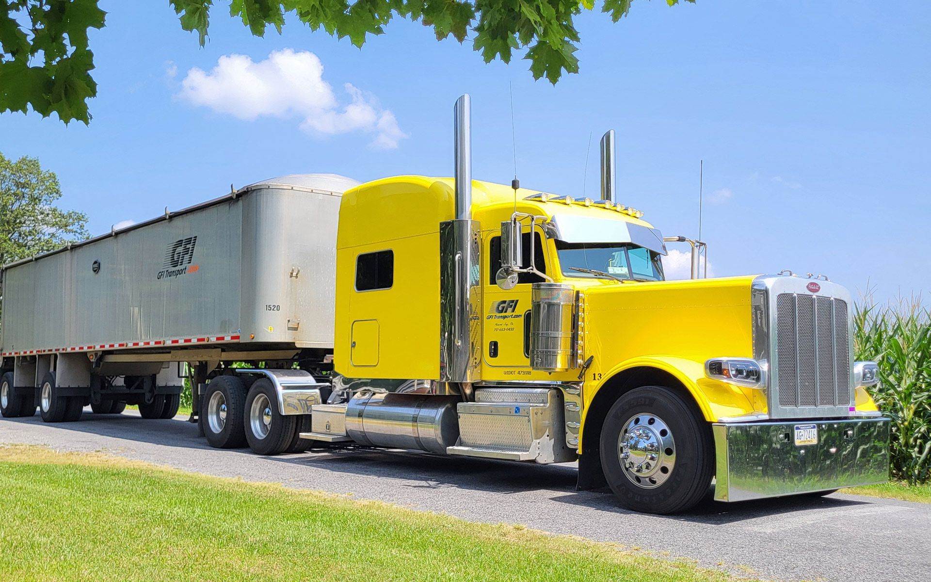 Bright yellow semi-truck parked on grass, hauling a long silver trailer under a blue sky.