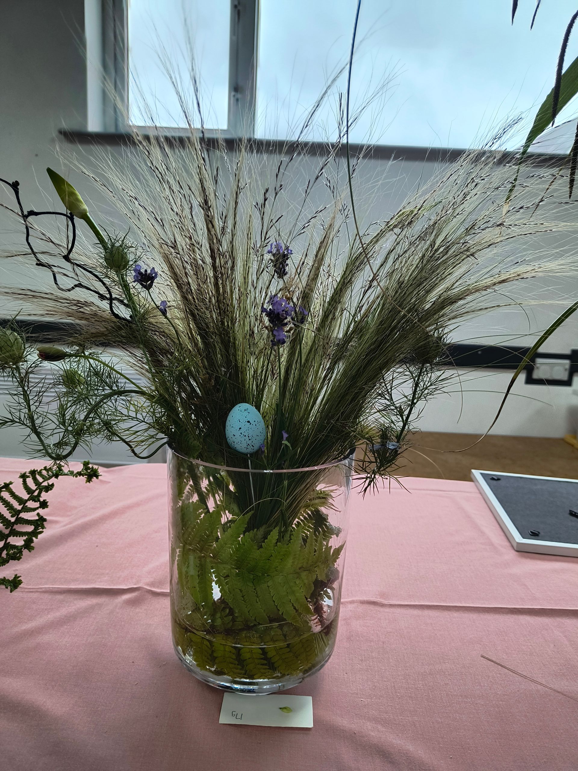 An arrangement of grasses, fens and other plants in a glass with a blue egg