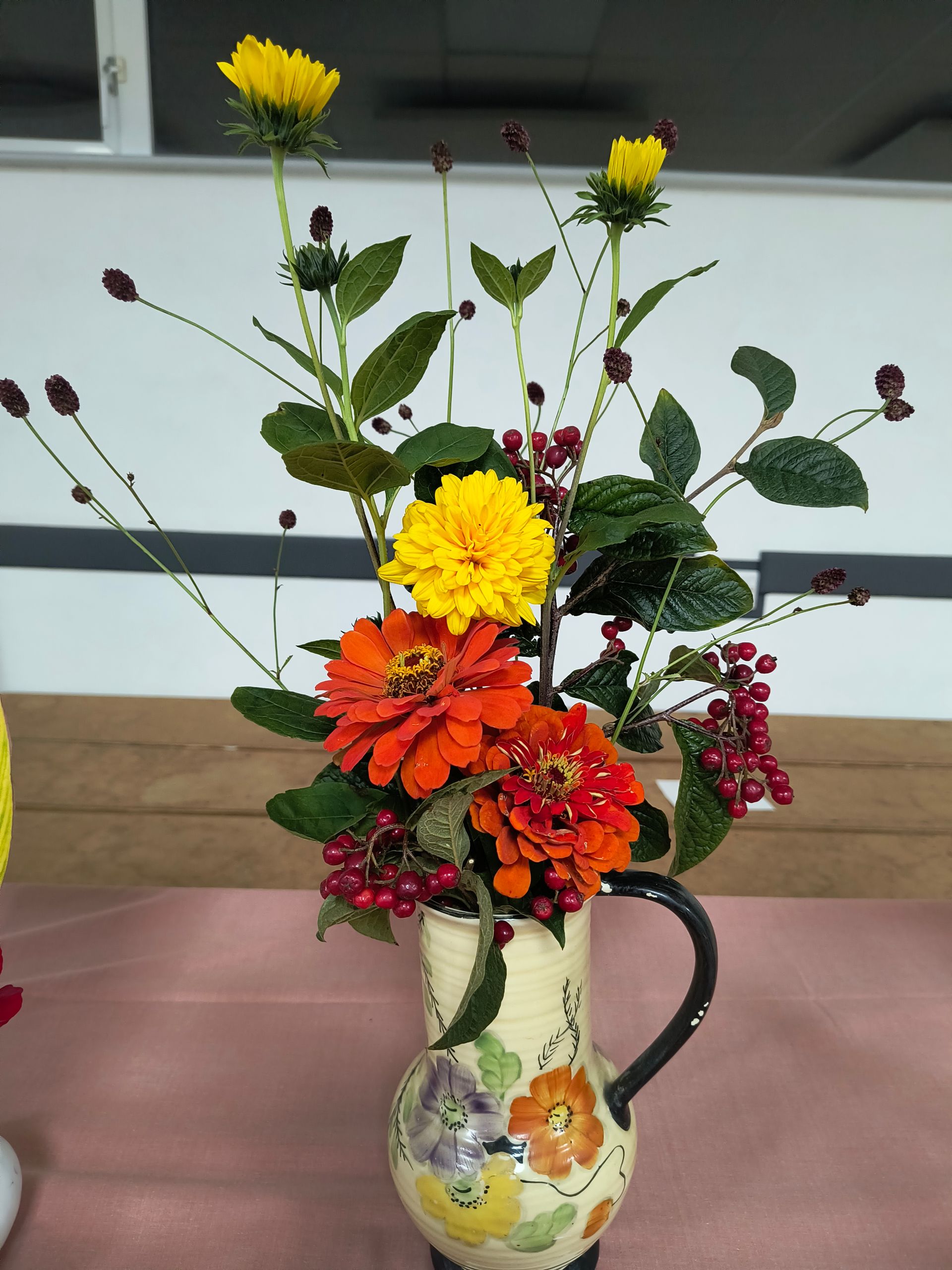 Photo shows orange, yellow and red flowers with foliage and red berries arranged in an jug
