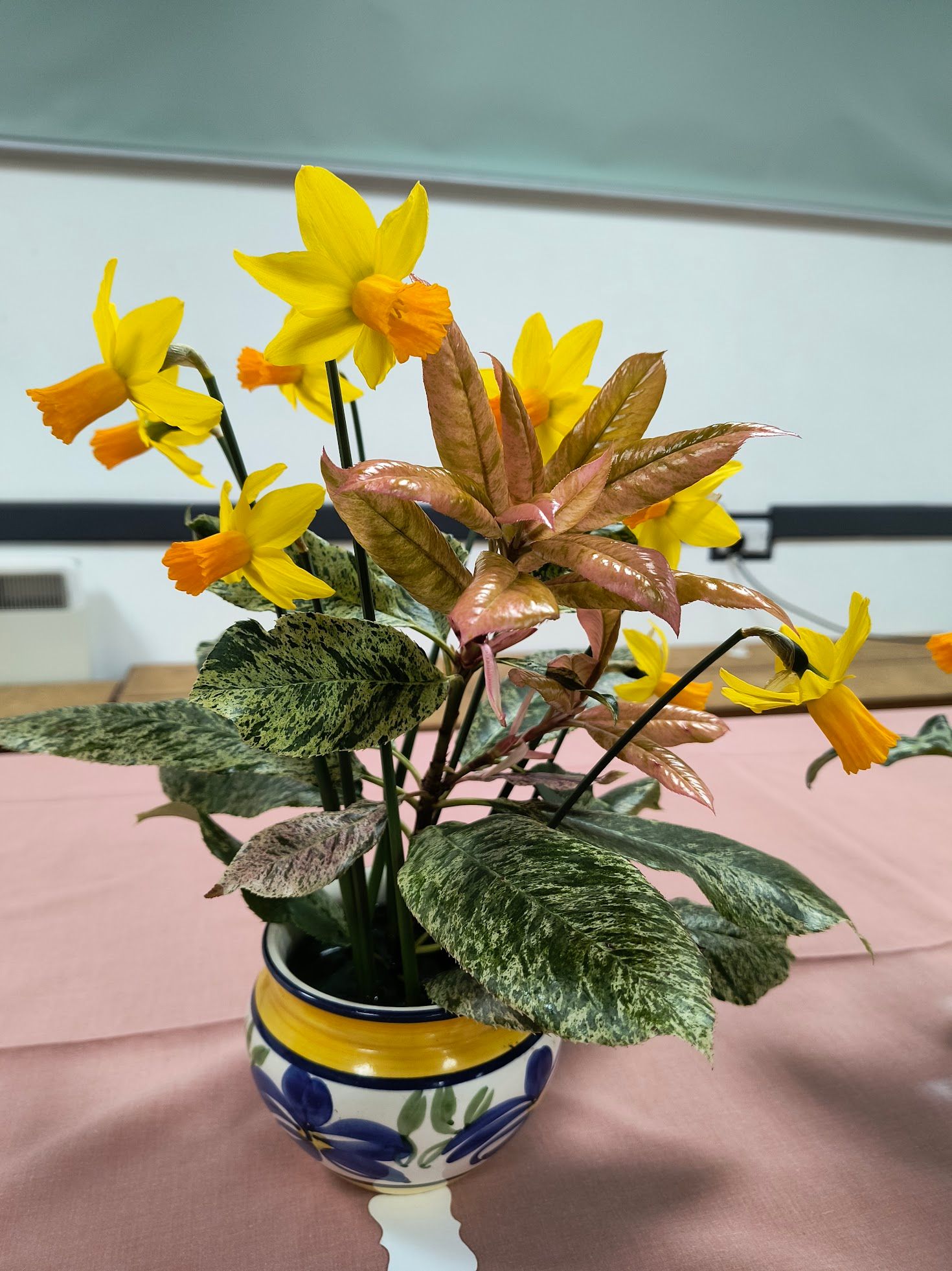 Photo shows an arrangement of deep yellow daffodils and various foliage in a ceramic vase