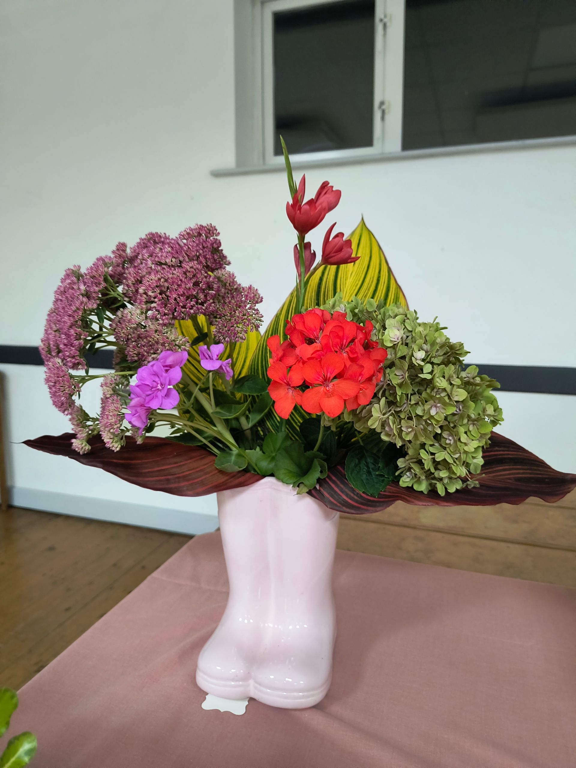 Photo shows pink and red flowers and green hydrangea head with canna leaves in a pink Wellington Boot shaped vase