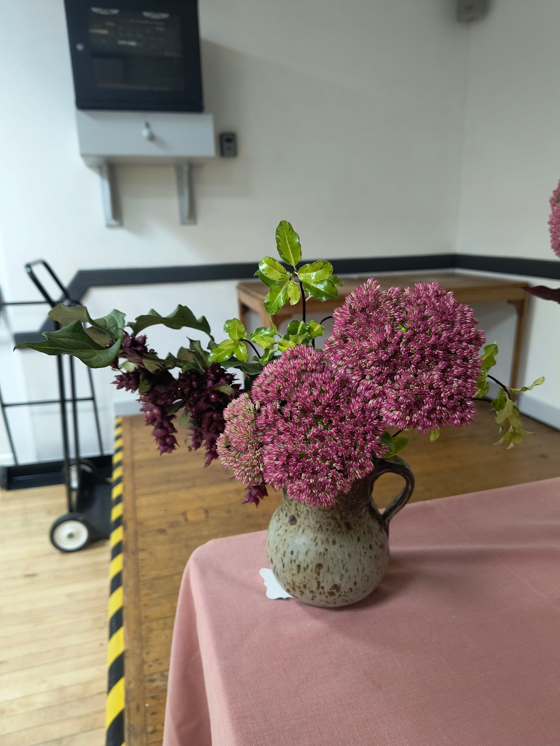 Photo shows dark pink flowers and foliage in a grey jug